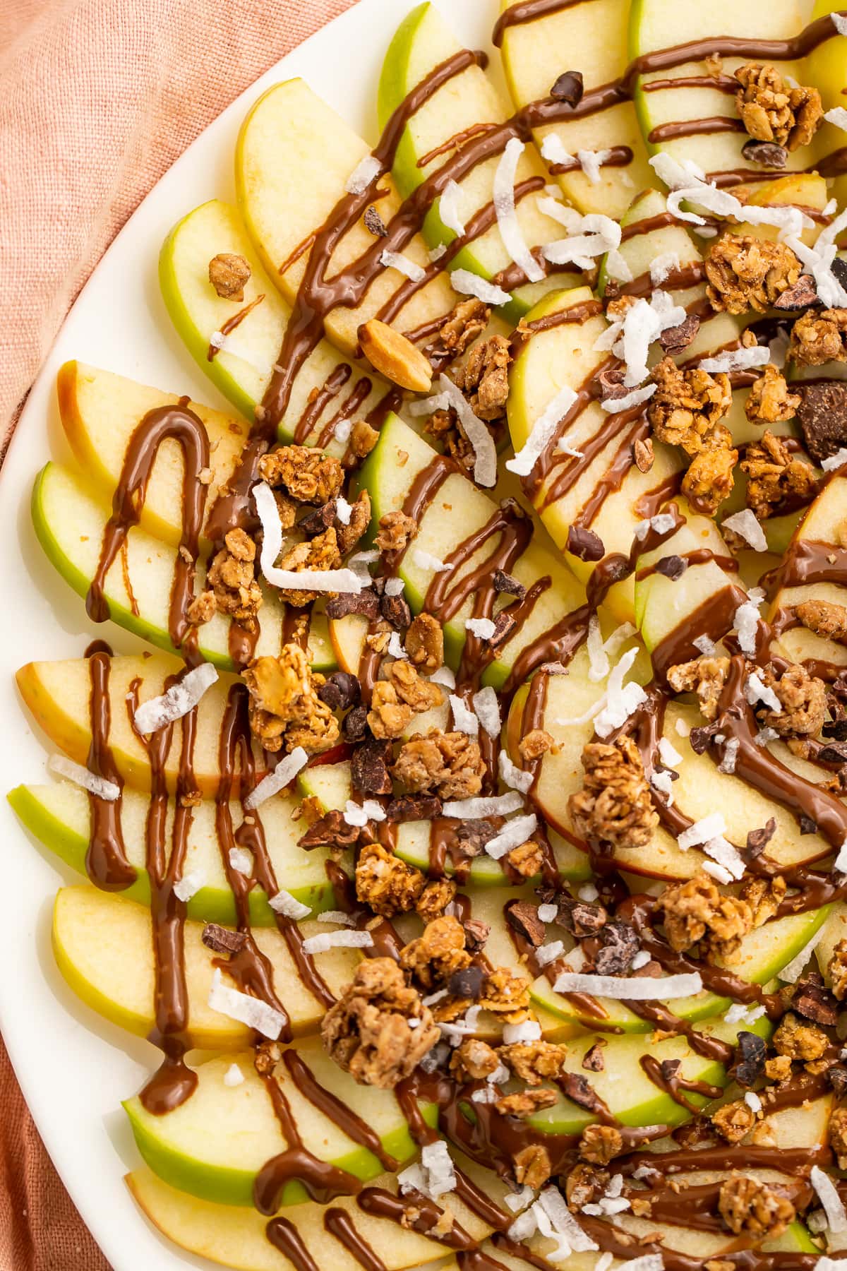 Overhead, top-down look at a plate of green and red apple slices topped with swirls of chocolate syrup and granola.