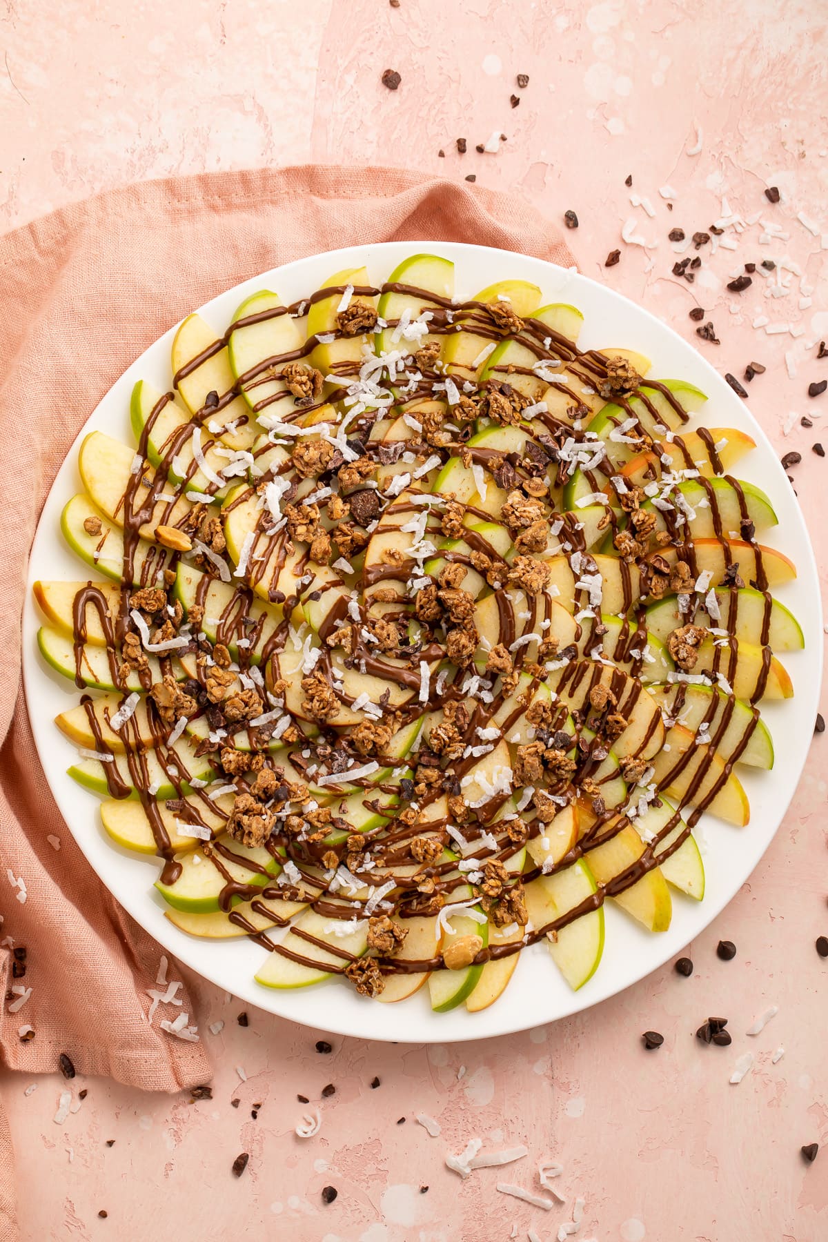 Overhead, top-down look at a plate of green and red apple slices topped with swirls of chocolate syrup and granola.