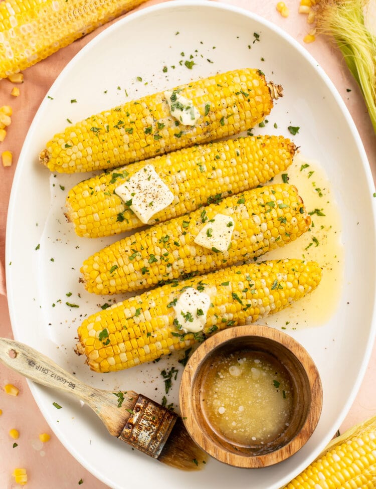 4 ears of air fryer corn on the cob arranged on a white oval platter next to a ramekin of melted butter.