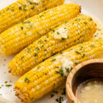 A close-up of 4 ears of air fryer corn on the cob topped with pats of butter and fresh parsley to garnish.