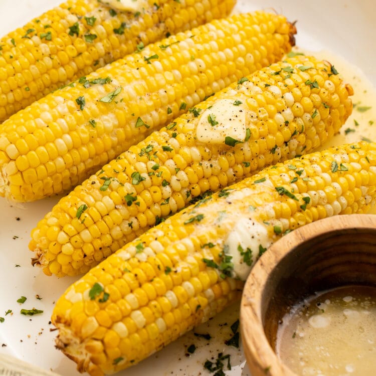 A close-up of 4 ears of air fryer corn on the cob topped with pats of butter and fresh parsley to garnish.