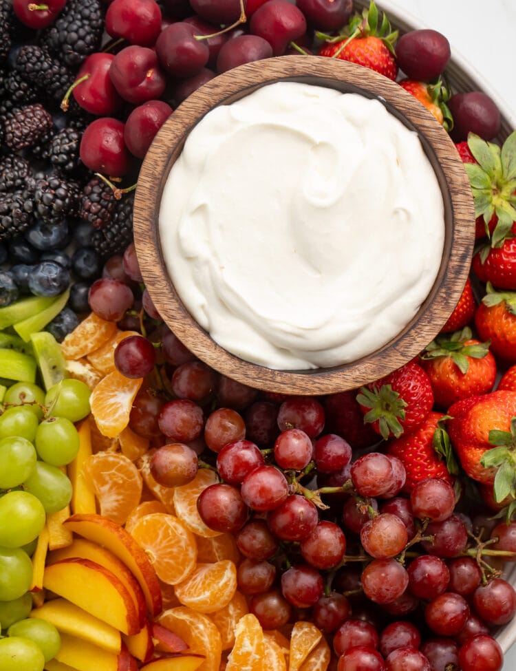 A small wooden bowl containing a white, whipped cream cheese dip on a platter surrounded by a rainbow of fruit.