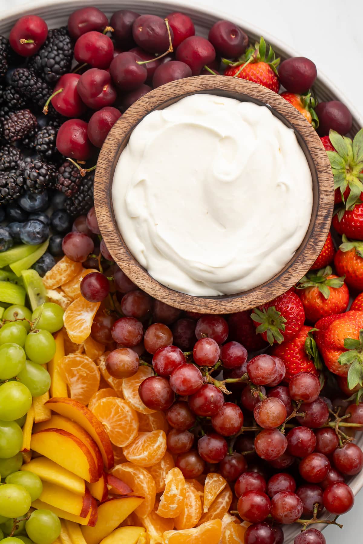 A small wooden bowl containing a white, whipped cream cheese dip on a platter surrounded by a rainbow of fruit.