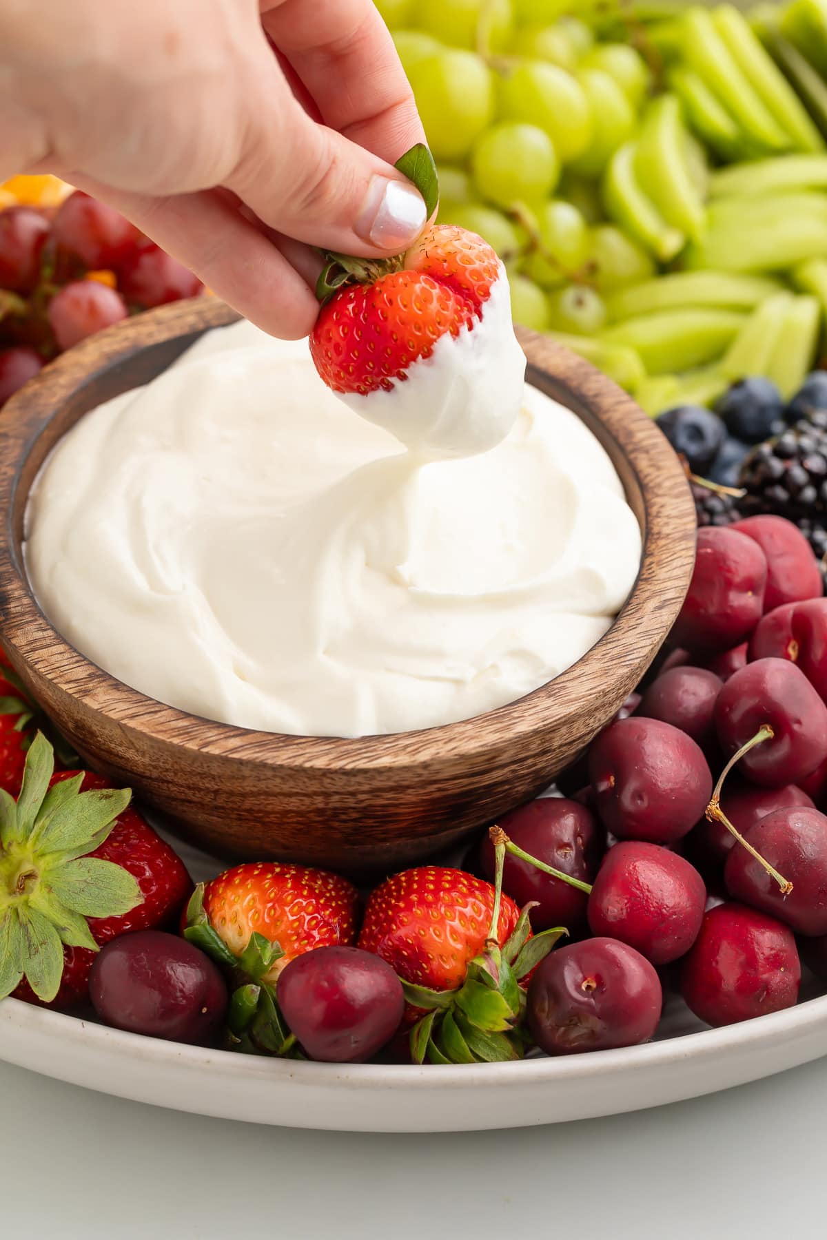 A white woman's hand holding a fresh strawberry as the woman dips it into a bowl of whipped cream cheese.
