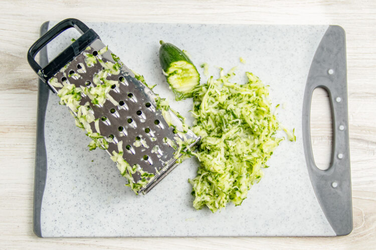 Green, unpeeled cucumber grated onto a cutting board with a box grater.