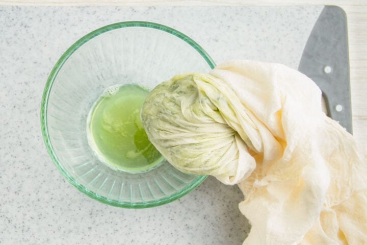 Pale green liquid in a small glass bowl next to grated cucumber in a cheesecloth.