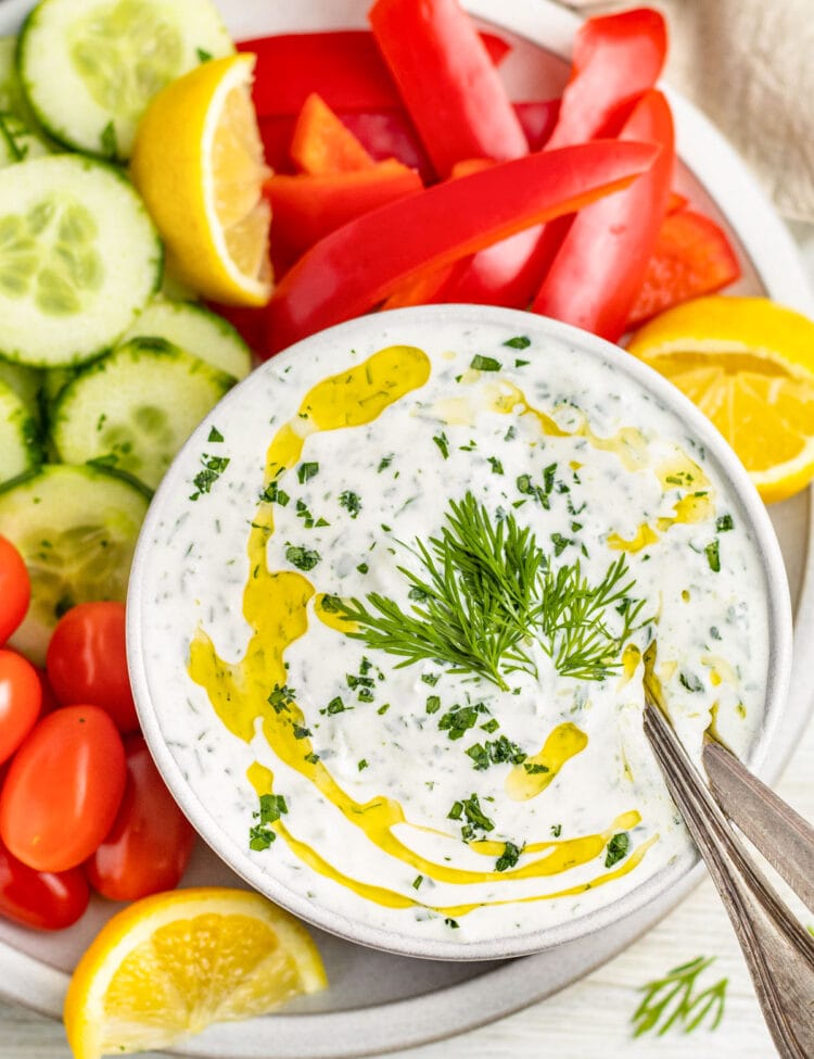 A bowl of creamy Whole30 tzatziki dotted with green fresh herbs and topped with a drizzle of olive oil, surrounded by cucumber slices, grape tomatoes, and bell pepper slices.
