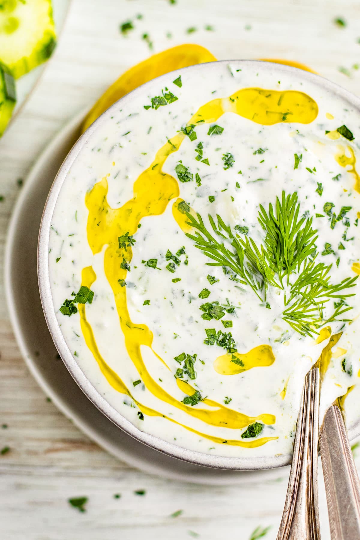 Close-up of a white bowl containing white Whole30 tzatziki dotted with bright green fresh herbs and topped with a drizzle of rich yellow olive oil.