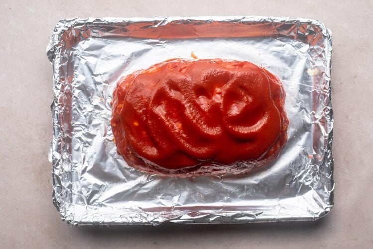 Unbaked meatloaf topped with a bright red ketchup glaze on a baking sheet lined with aluminum foil.