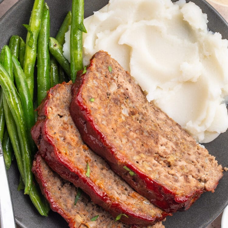 Three slices of gluten-free meatloaf topped with ketchup and plated with mashed potatoes and fresh green beans.