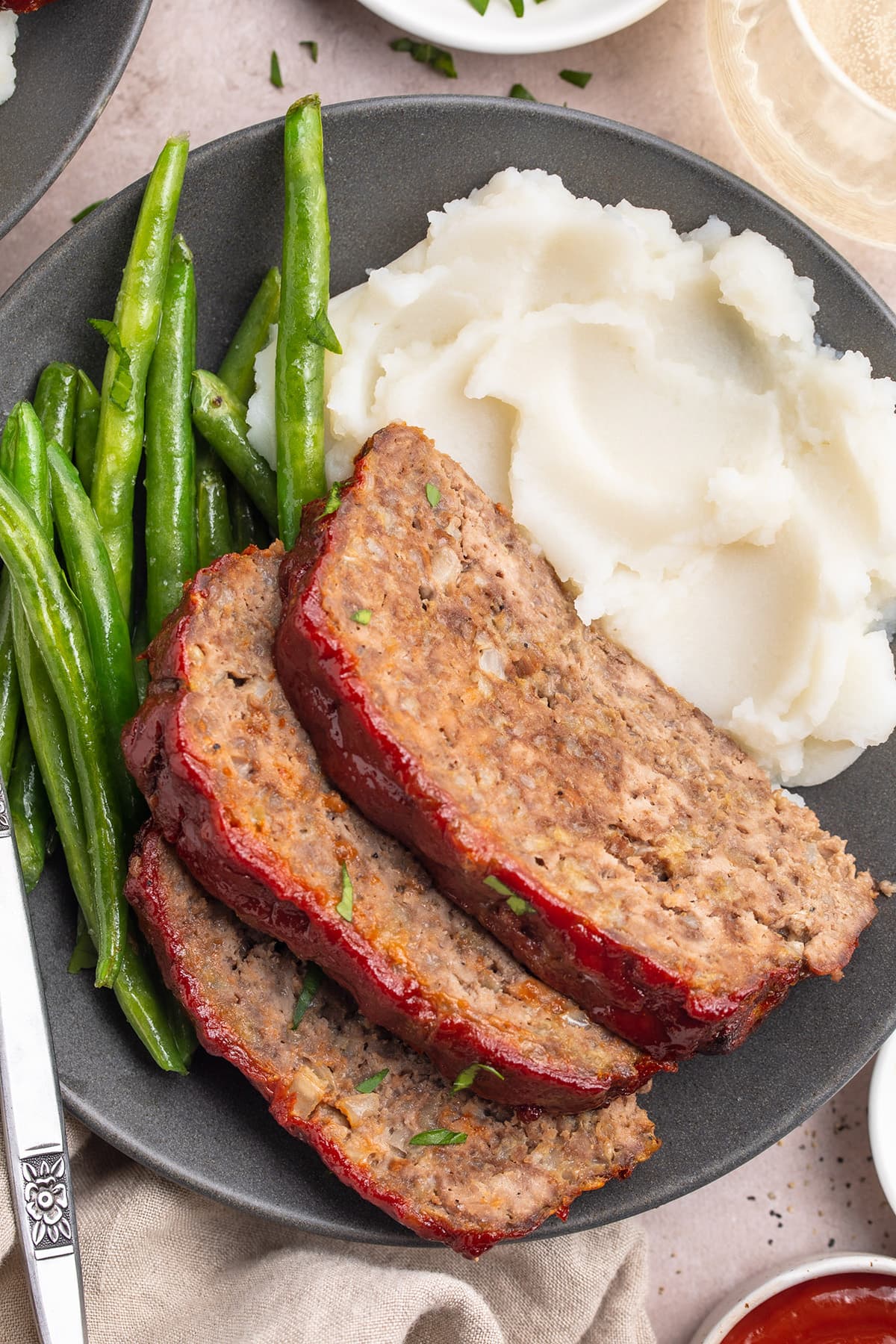 Three slices of gluten-free meatloaf topped with ketchup and plated with mashed potatoes and fresh green beans.