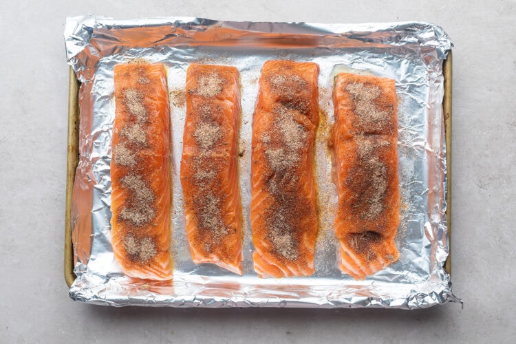 Four salmon fillets on a foil-lined baking sheet, topped with neutral oil and jerk seasoning.