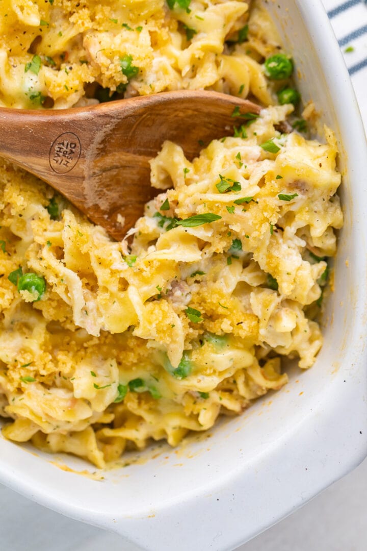 A large oval casserole dish holding tuna noodle casserole, with a large wooden spoon resting in the dish.
