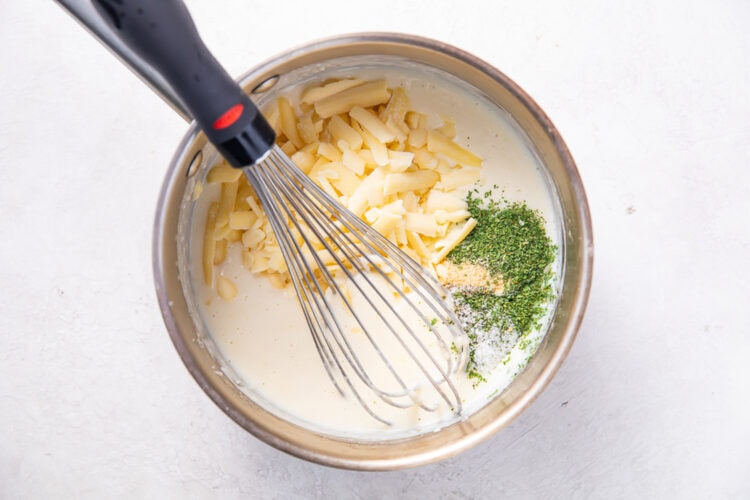 Seasonings added to cream sauce base in a silver saucepan with a silver wire whisk.