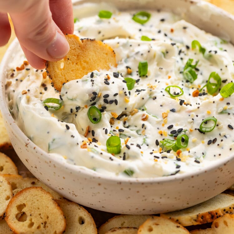 A white woman's hand dipping a bagel chip into a bowl of Everything Bagel dip surrounded by other bagel chips.