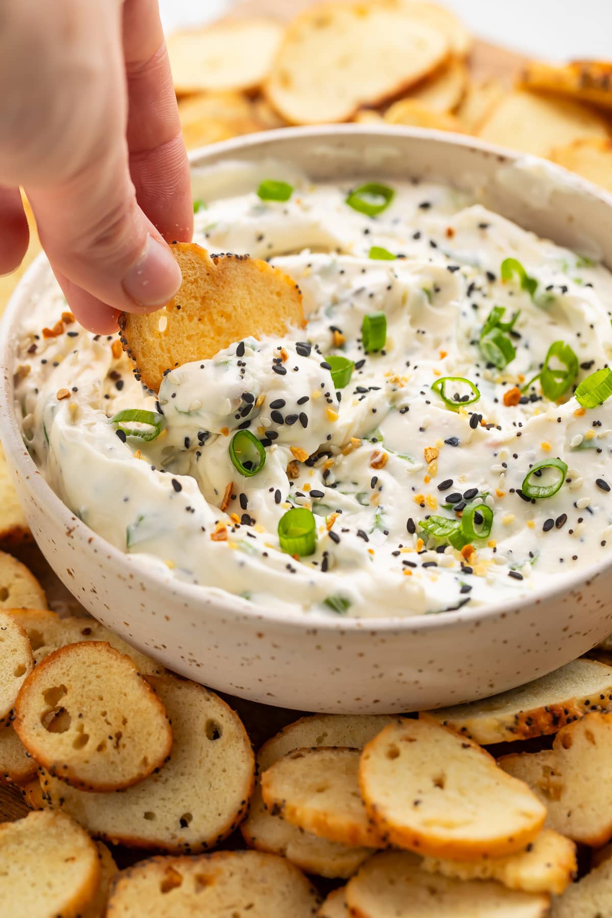 A white woman's hand dipping a bagel chip into a bowl of Everything Bagel dip surrounded by other bagel chips.