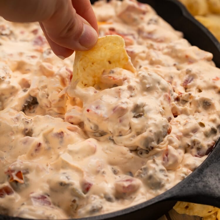 A white woman's hand dipping a scoop-shaped tortilla chip into a skillet full of cream cheese sausage dip.