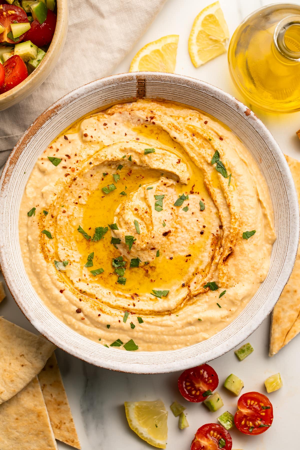 Overhead view of a white bowl holding hummus made without tahini, swirled with olive oil and garnished with herbs.