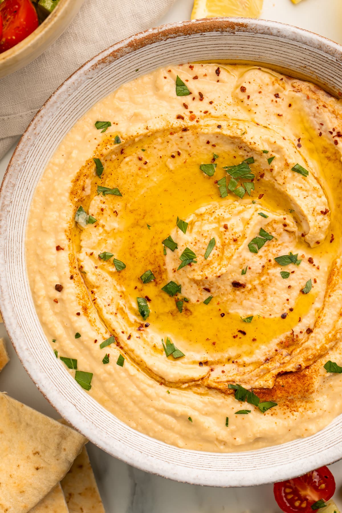 Close-up overhead view of a white bowl holding hummus made without tahini, swirled with olive oil and garnished with herbs.