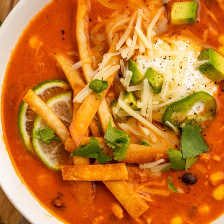 Close-up photo of a large white bowl holding deep red chicken tortilla soup, made in an Instant Pot, topped with tortilla strips, cheese, avocado, and cilantro.