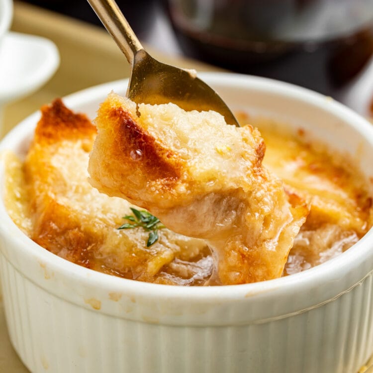A spoon lifting a piece of French bread off the top of a ramekin holding Instant Pot French onion soup.