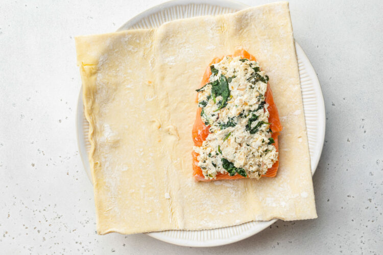 A sheet of puff pastry topped with a salmon fillet and cream cheese spinach filling on a white plate.