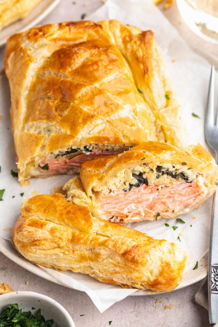 Salmon wellington, sliced and plated on a white round plate, with one slice placed on its side to show the salmon filling.