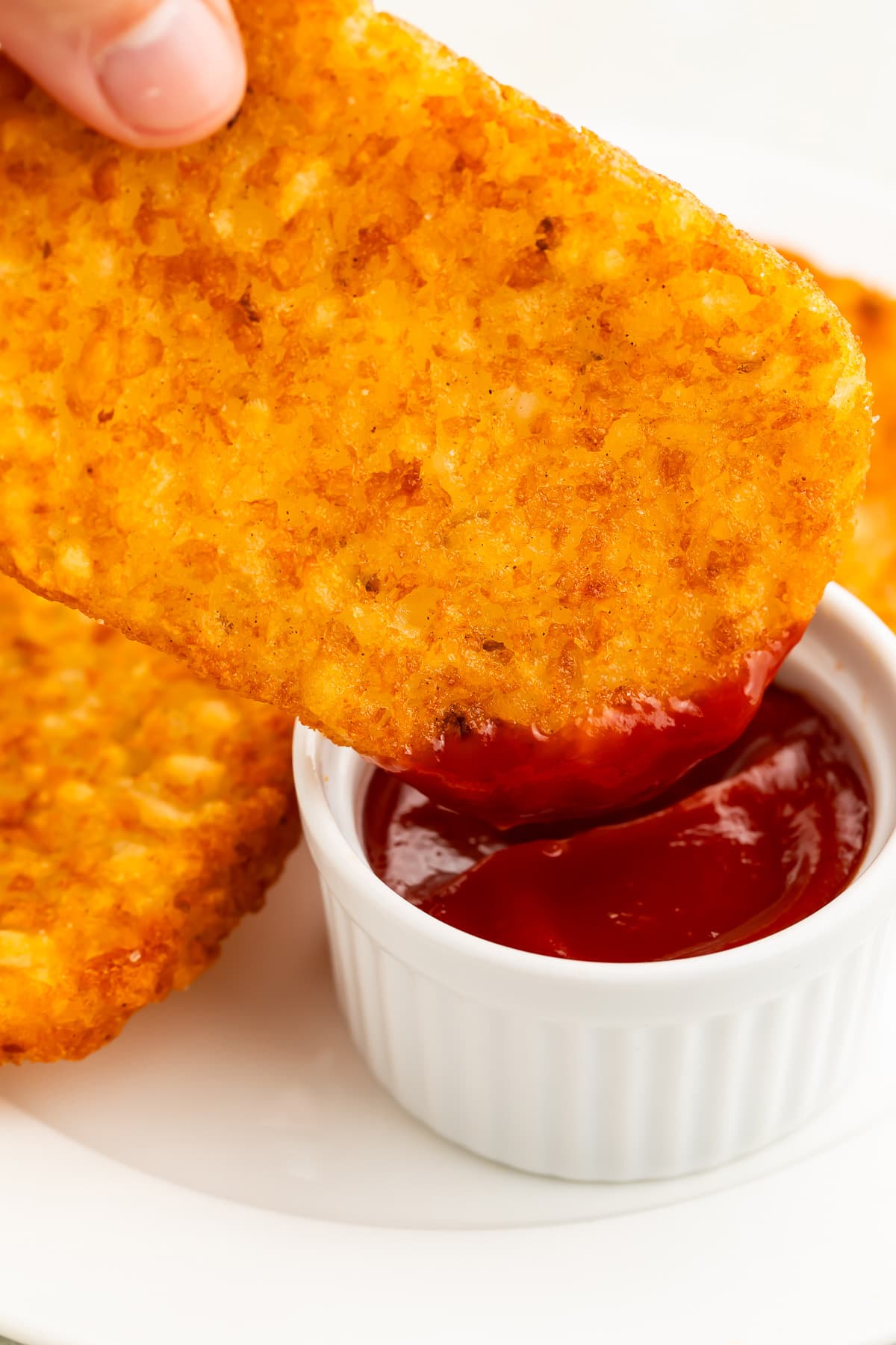 1 of 4 frozen hash brown patties, cooked in the air fryer, being dipped into a small ramekin of red ketchup.