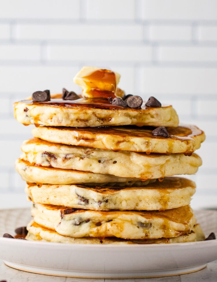 A stack of chocolate chip pancakes topped with chocolate chips, two pats of butter, and plenty of maple syrup.