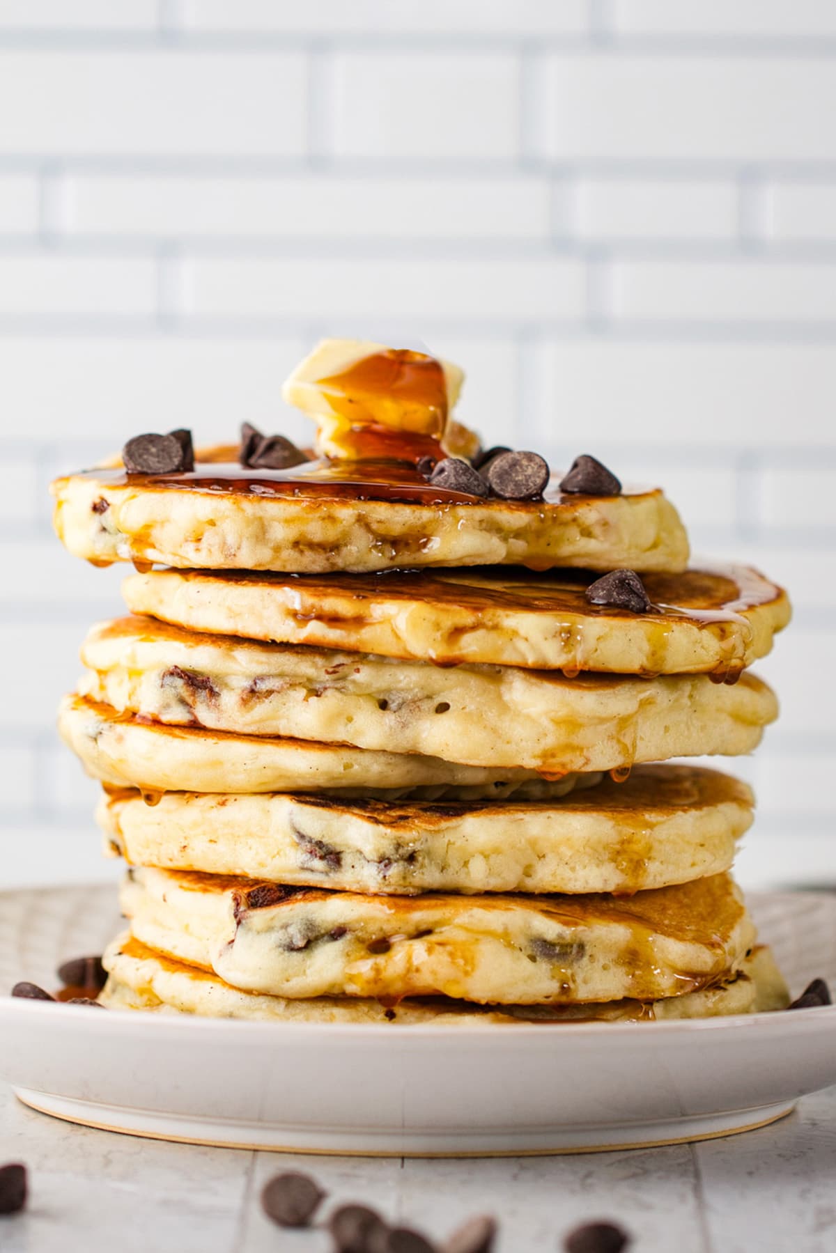 A stack of chocolate chip pancakes topped with chocolate chips, two pats of butter, and plenty of maple syrup.