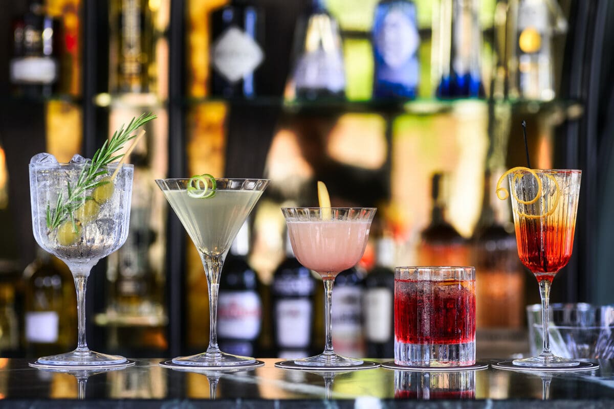 5 prepared cocktails on a bar in front of a wall of alcohol bottles.