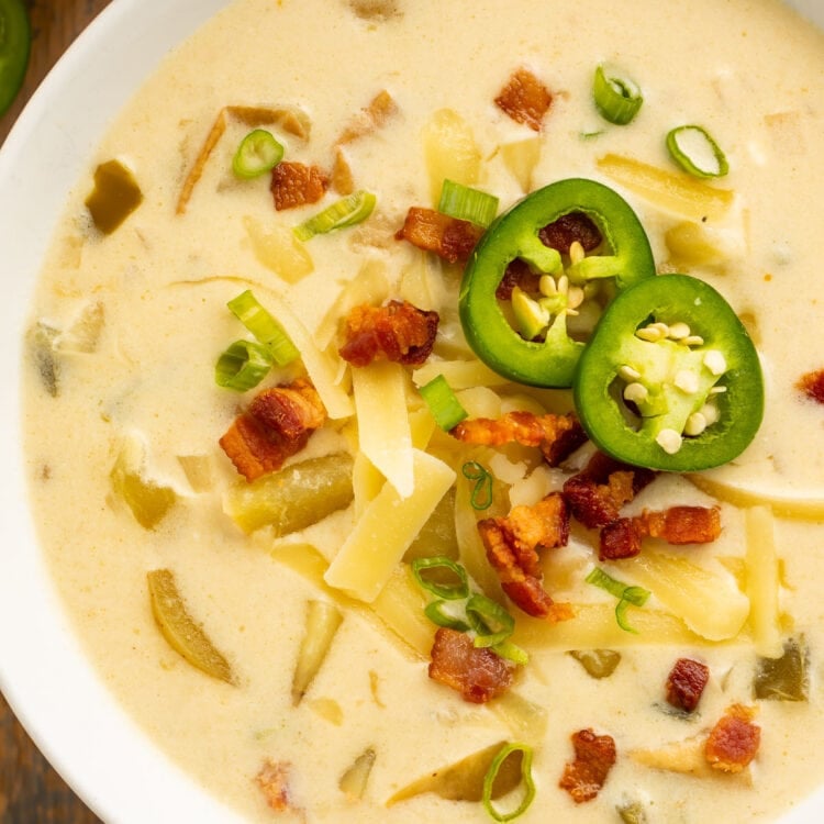 Top-down view of a large white bowl holding pale yellow, creamy jalapeño soup, topped with sliced jalapeños, diced bacon, and shredded cheese.