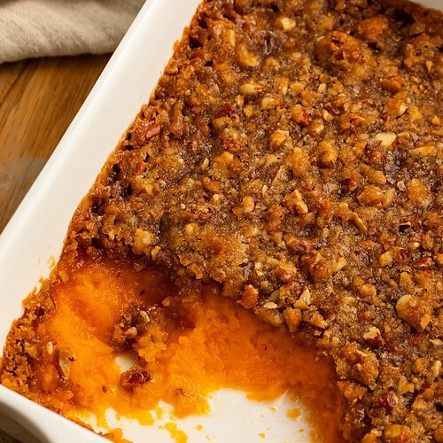 Close up of a sweet potato casserole with a spoonful missing, and a crunchy pecan praline topping.