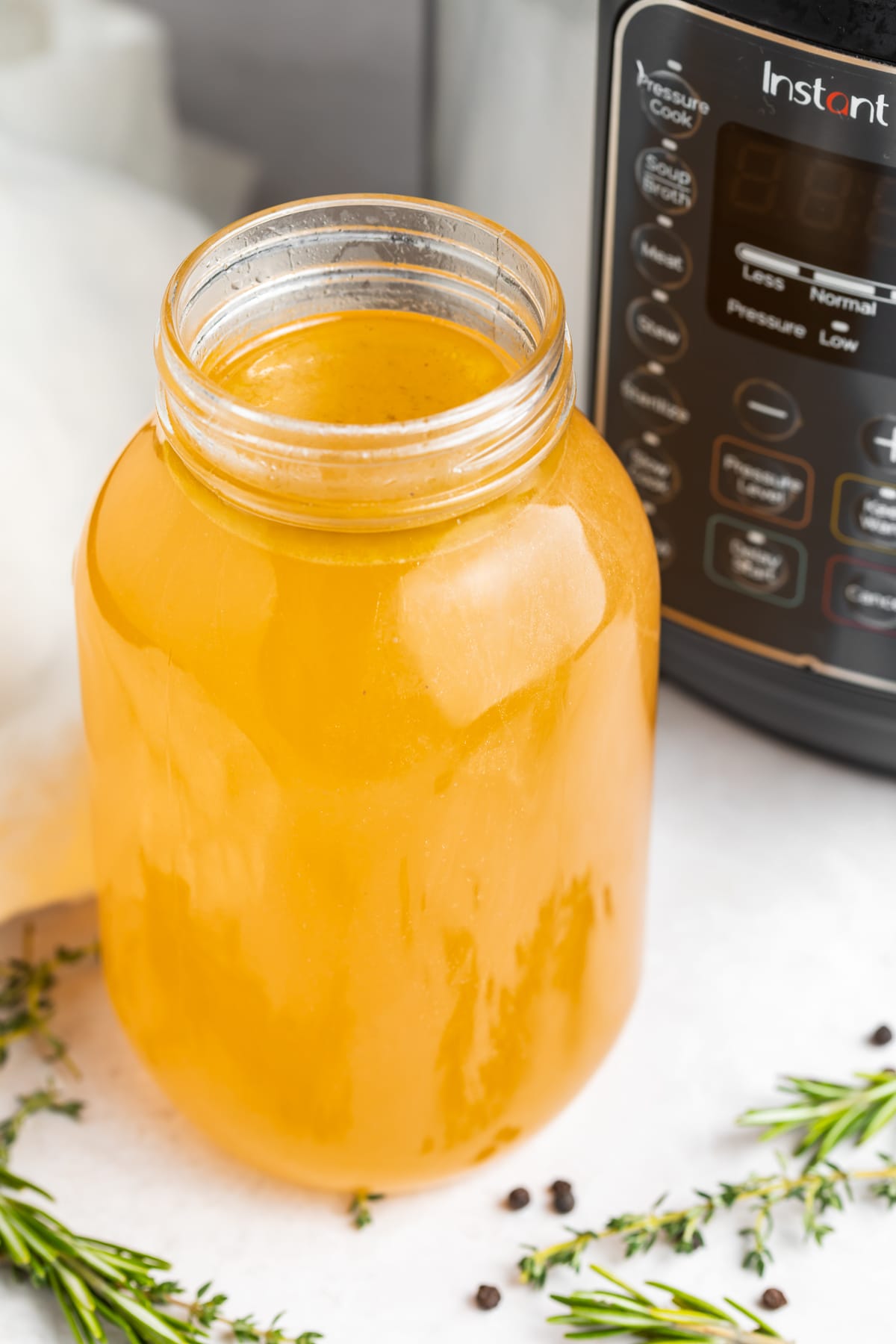 A mason jar of golden yellow turkey stock resting in front of an Instant Pot surrounded by fresh herbs.