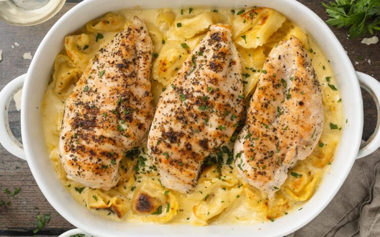 Grilled chicken breasts on a bed of asiago tortelloni with alfredo sauce in a casserole dish.