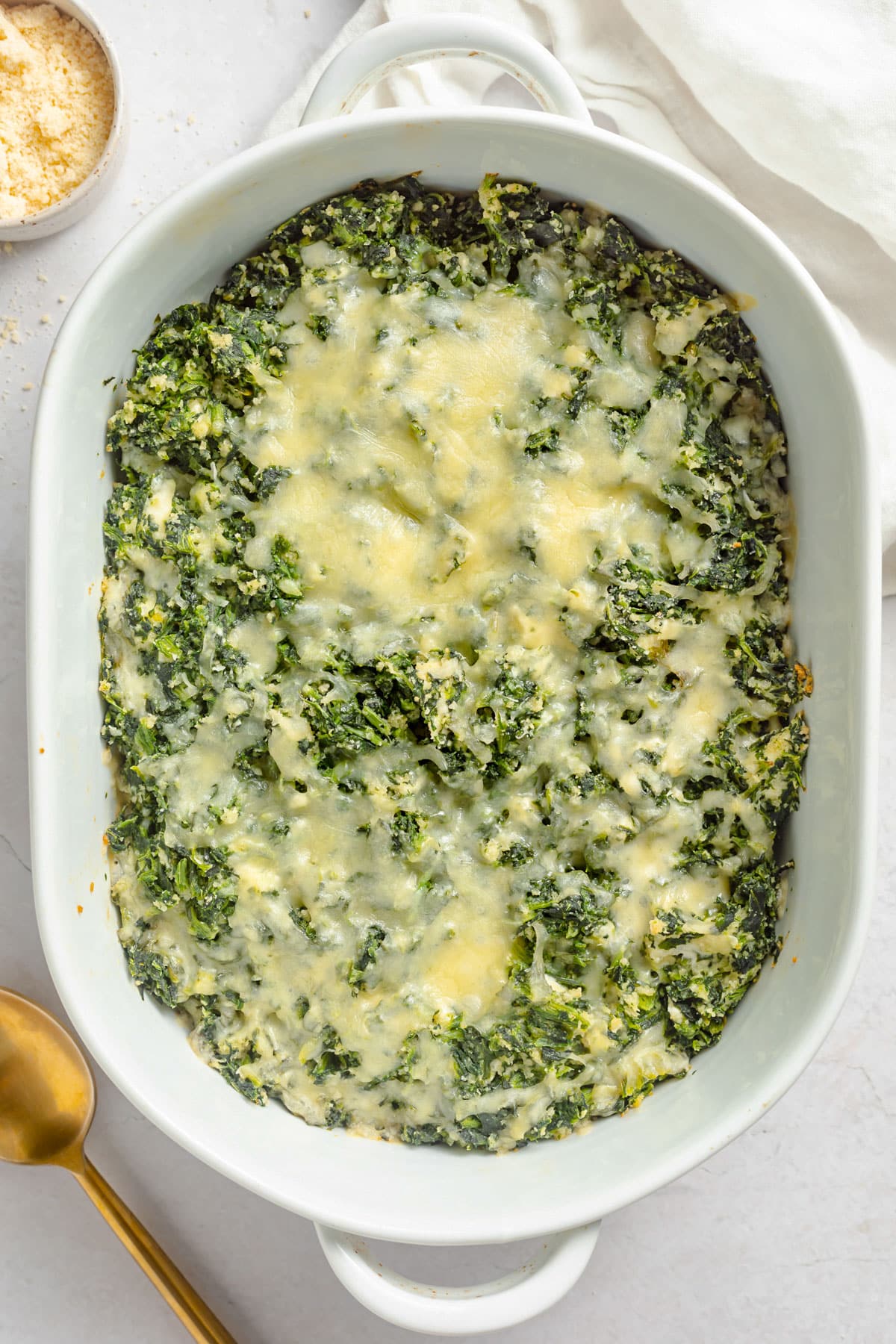 Top-down view of a large oval casserole dish holding a cheesy spinach casserole topped with parmesan.