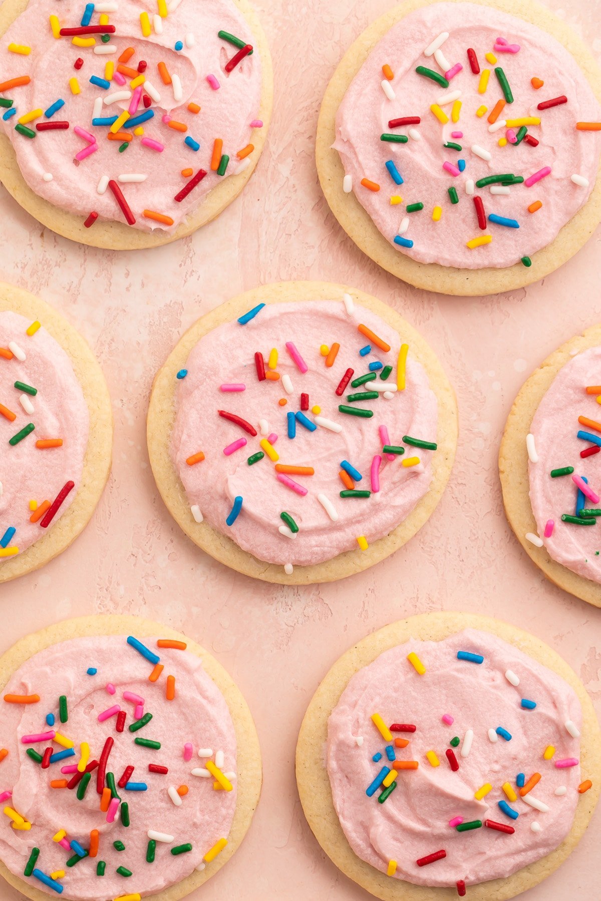 Rows of gluten free sugar cookies topped with pink buttercream frosting and rainbow jimmies on parchment paper.