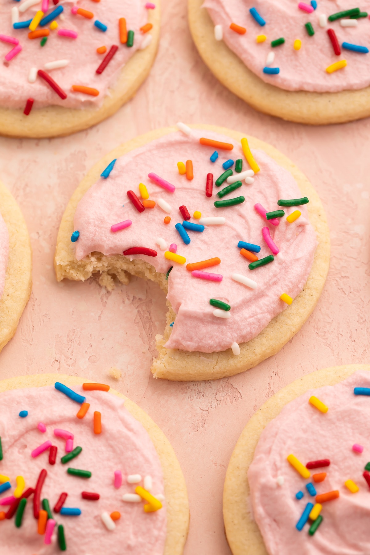 Rows of gluten free sugar cookies topped with pink buttercream frosting and rainbow jimmies on parchment paper. One bite is missing from the cookie in the center of the photo.