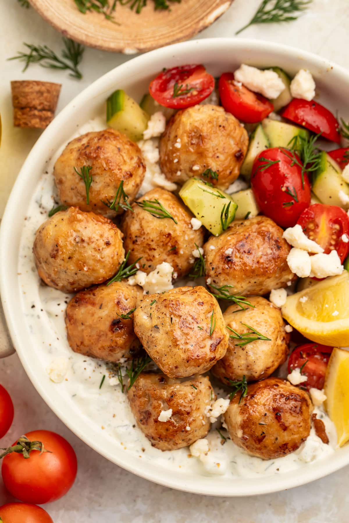 Top-down photo showing Greek chicken meatballs in a large bowl with rice, feta cheese, veggies, and herbs.