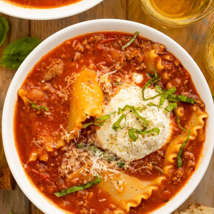 A bowl of lasagna soup on a wooden table next to a piece of bread and a glass of wine.