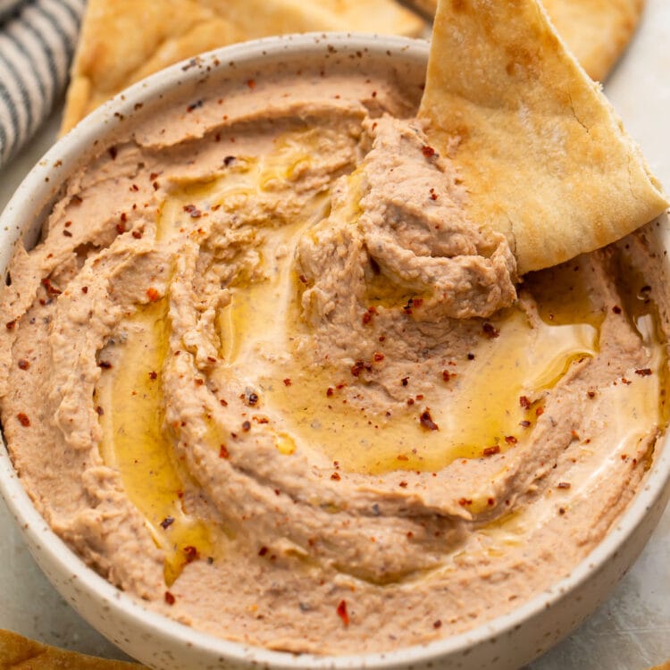 A triangular pita chip resting in a bowl of black eyed pea hummus, with a scoop of hummus on the chip.