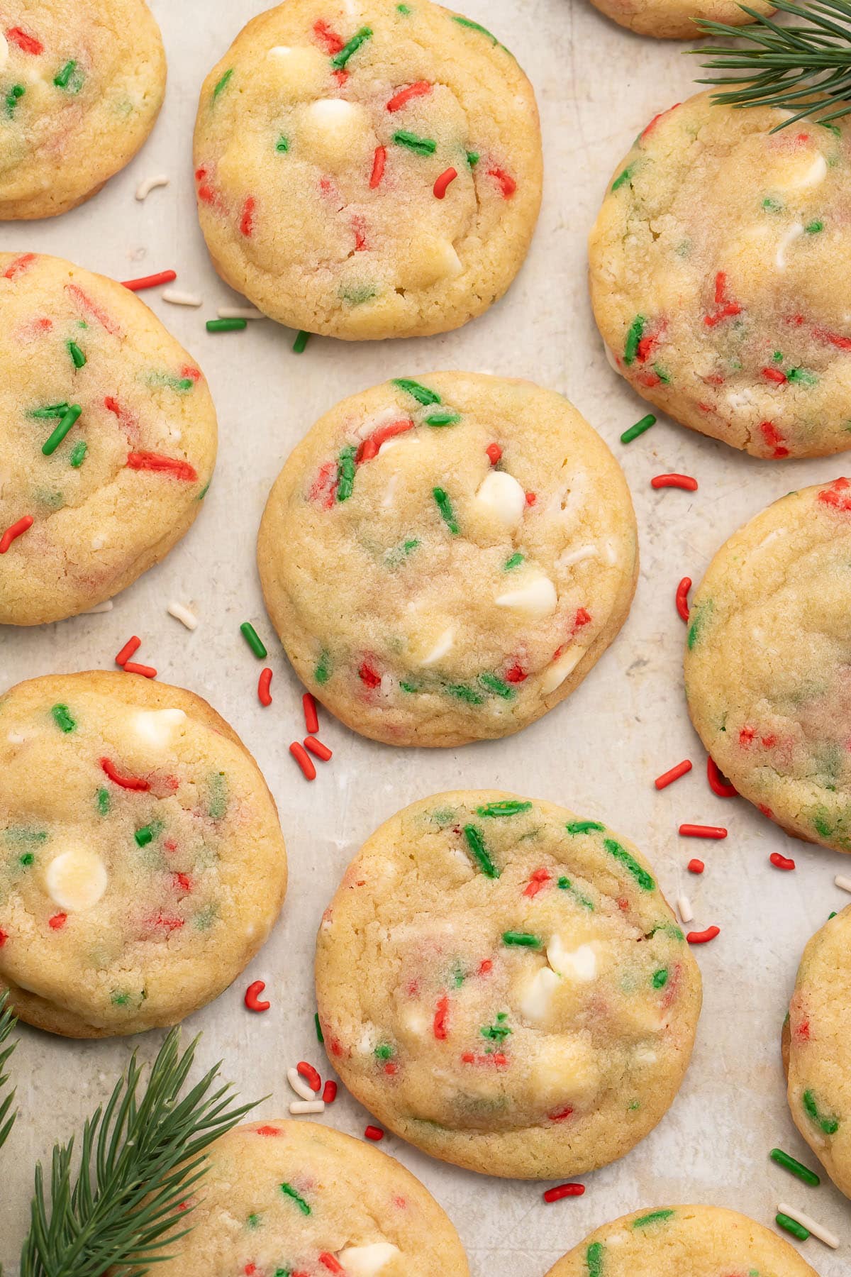 Rows of Christmas confetti cookies with red, white, and green sprinkles on parchment paper, with sprigs of rosemary resembling Christmas tree branches at the corners of the image.