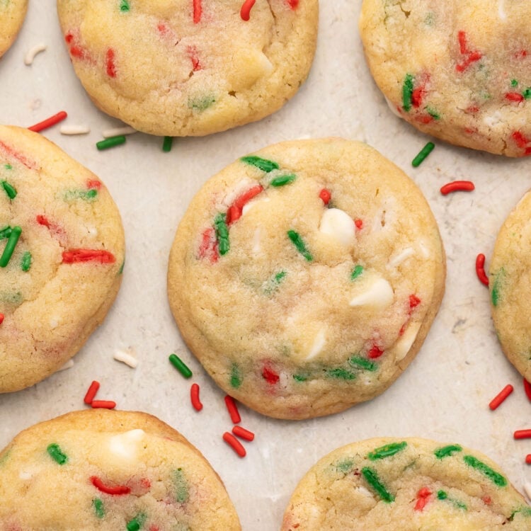 Rows of Christmas confetti cookies with red, white, and green sprinkles on parchment paper, with sprigs of rosemary resembling Christmas tree branches at the corners of the image.