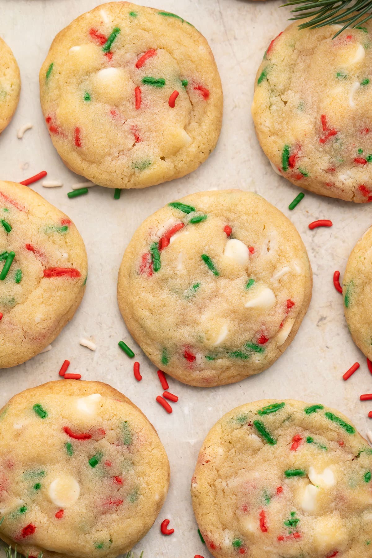 Rows of Christmas confetti cookies with red, white, and green sprinkles on parchment paper, with sprigs of rosemary resembling Christmas tree branches at the corners of the image.