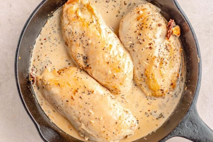 Three large seared chicken breasts smothered in cream sauce in a large cast-iron skillet.