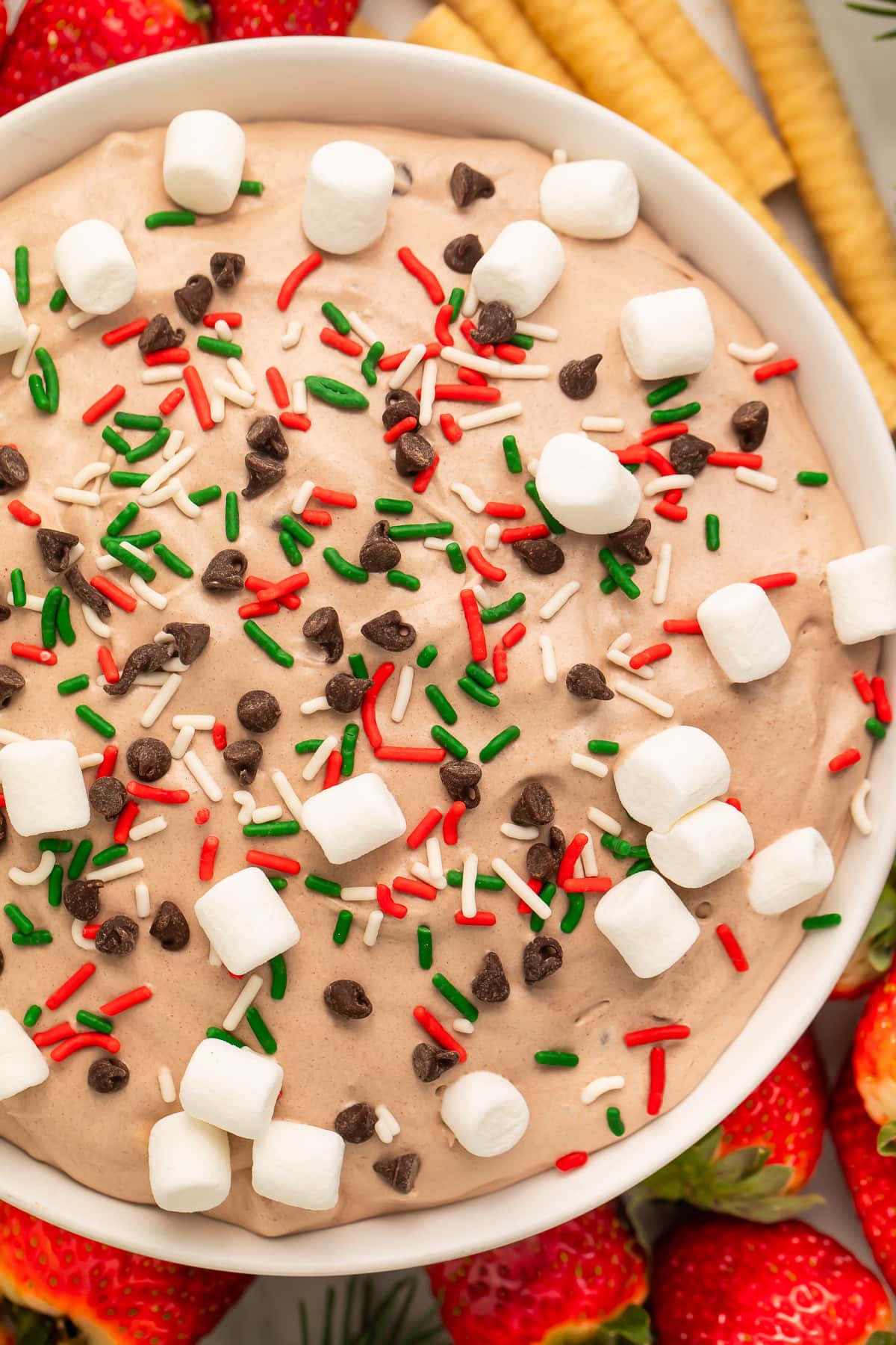 Top-down photo of hot cocoa dip in a large white bowl. The dip is topped with mini marshmallows, holiday sprinkles, and mini chocolate chips.