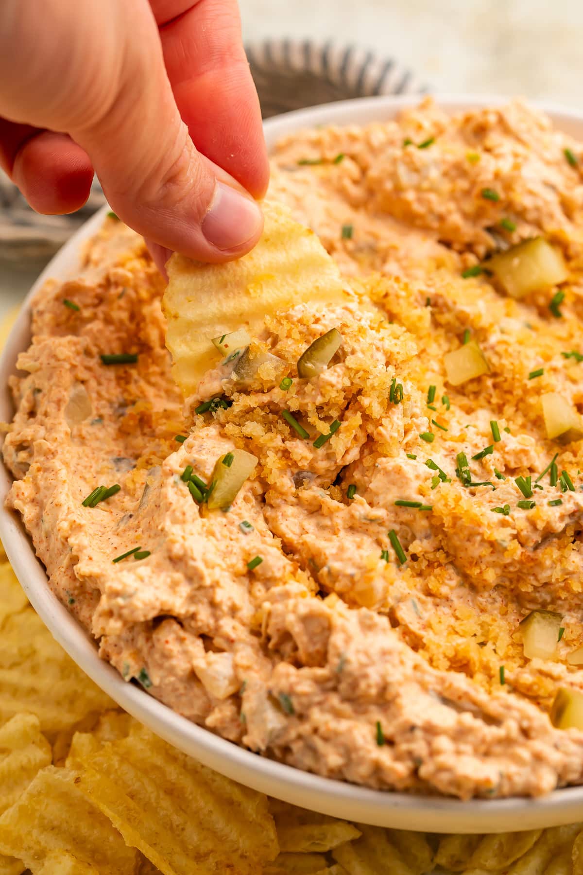 A white woman's hand holds a ridged potato chip, using it to scoop Nashville hot chicken dip out of a large bowl.