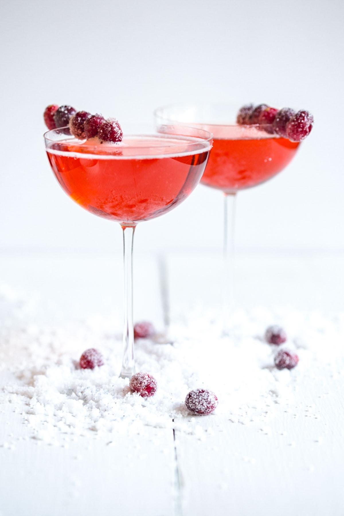 Two cocktail glasses holding bright red cranberry-elderflower champagne sparklers on a white table with sugared cranberries.