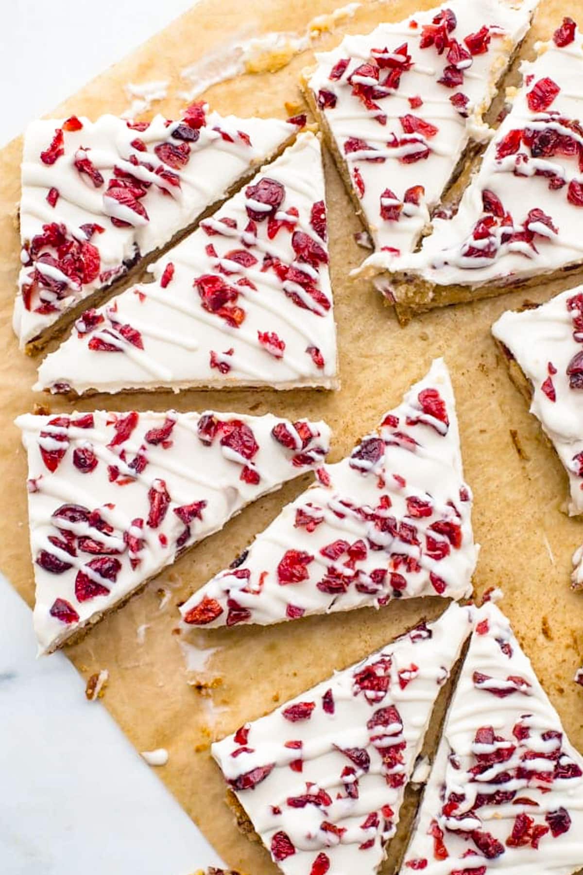 Overhead photo showing triangular cranberry bliss bars spread on a counter, partially on a piece of parchment paper.