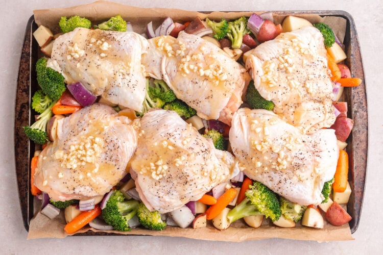 Chicken thighs topped with garlic butter sauce arranged atop a bed of vegetables on a sheet pan.